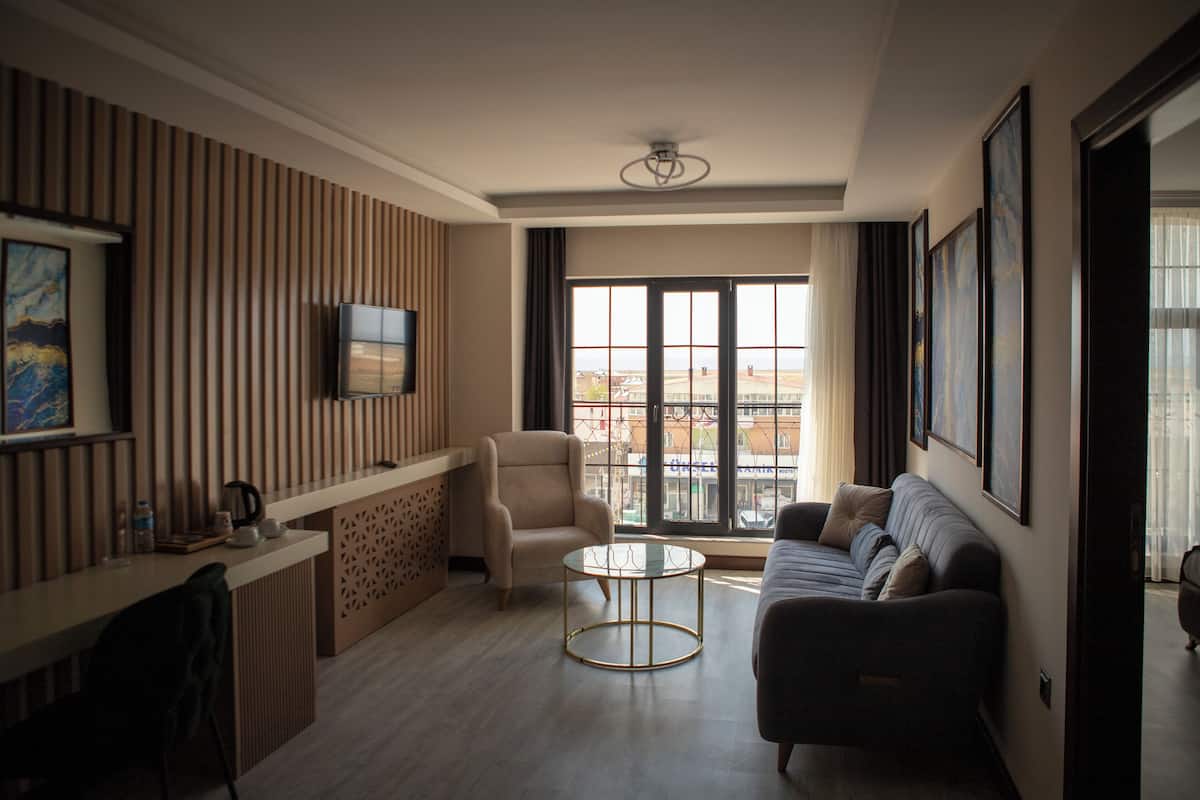 A modern living room with a sofa, armchair, and a small coffee table. There is a TV mounted on the wall and a window with a view of buildings.