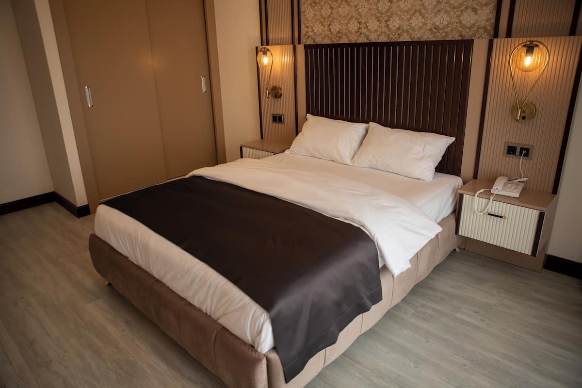 A modern hotel room with a large bed, wooden headboard, two bedside tables with lamps, and a built-in wardrobe.