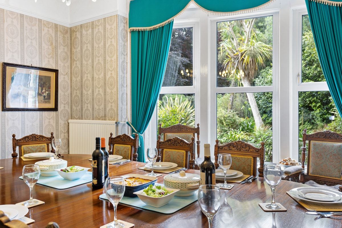 A dining room with a large table set for a meal, surrounded by ornate chairs, and large windows with curtains.