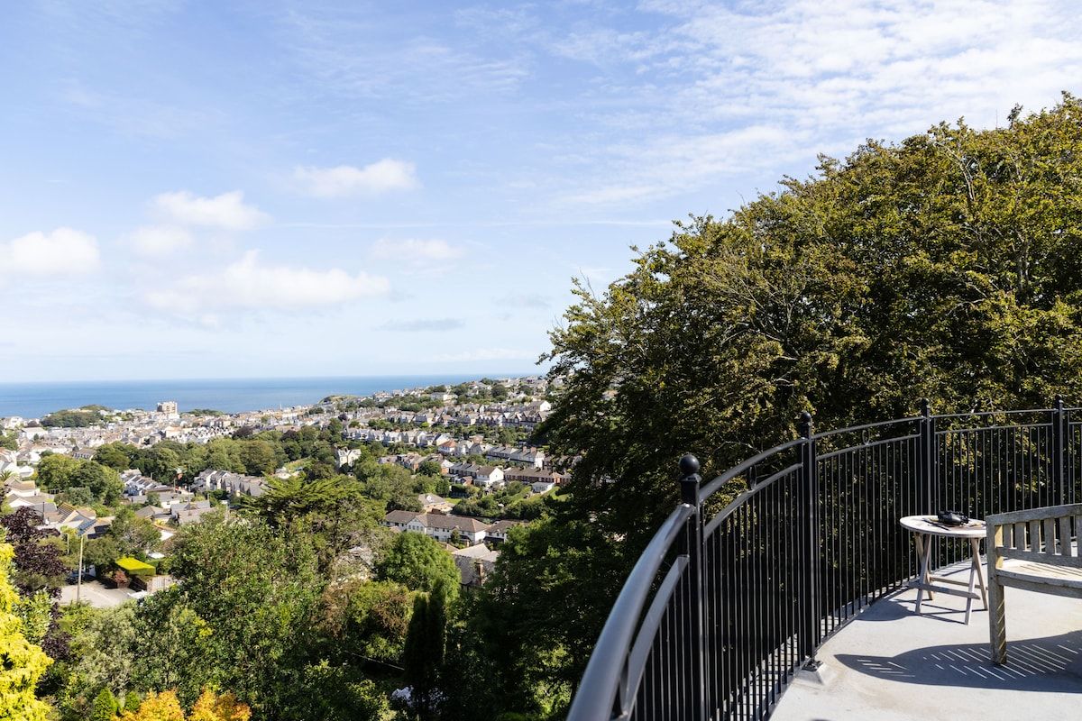 A balcony with a table and chair, overlooking a coastal town and the sea.