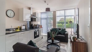 A modern kitchen with white cabinets, a black countertop, and a built-in oven. There is a dining area with a dark brown sofa and a round coffee table. A large window with vertical blinds allows natural light into the room. - Modern 1B Central Manchester + Free Parking (Salford)