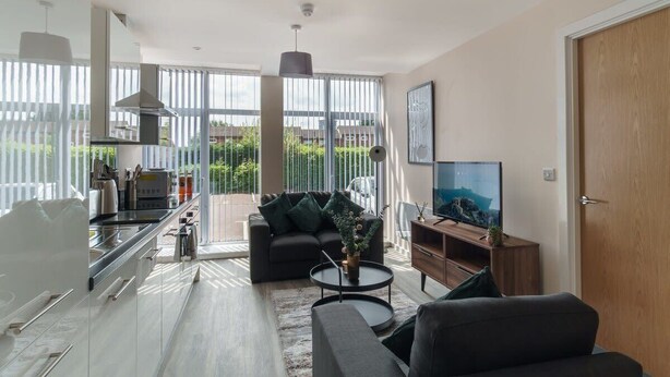 A modern living room with a sofa, coffee table, and TV. There is a kitchen area with a stove and cabinets. Large windows provide natural light and a view of greenery outside. - Modern 1B Central Manchester + Free Parking (Salford)