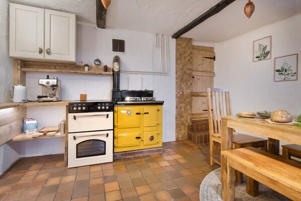 A kitchen with a stove, oven, and yellow cabinets. There is a wooden table with a chair, and a window with curtains. - The Gilpin Farmhouse, Lake's Escape | Ridgestone (Levens)