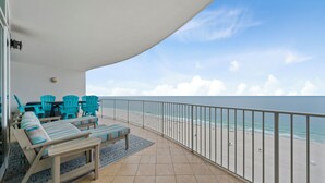 Uma varanda com vista para a praia, área de estar com cadeiras e sofá, e um corrimão.