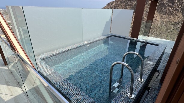 A glass-enclosed infinity pool with metal ladders, surrounded by a wooden deck and a glass railing, overlooking a mountainous landscape.