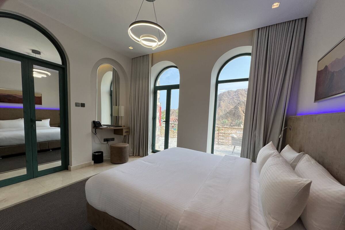 A modern hotel room with a large bed, a desk with a lamp, and a view of the mountains through the windows.