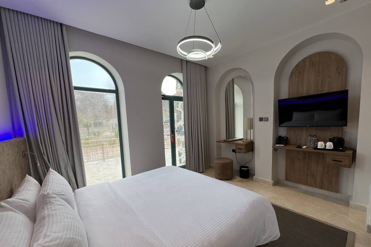 A modern hotel room with a large bed, a desk with a flat-screen TV, and a view of the outdoors.