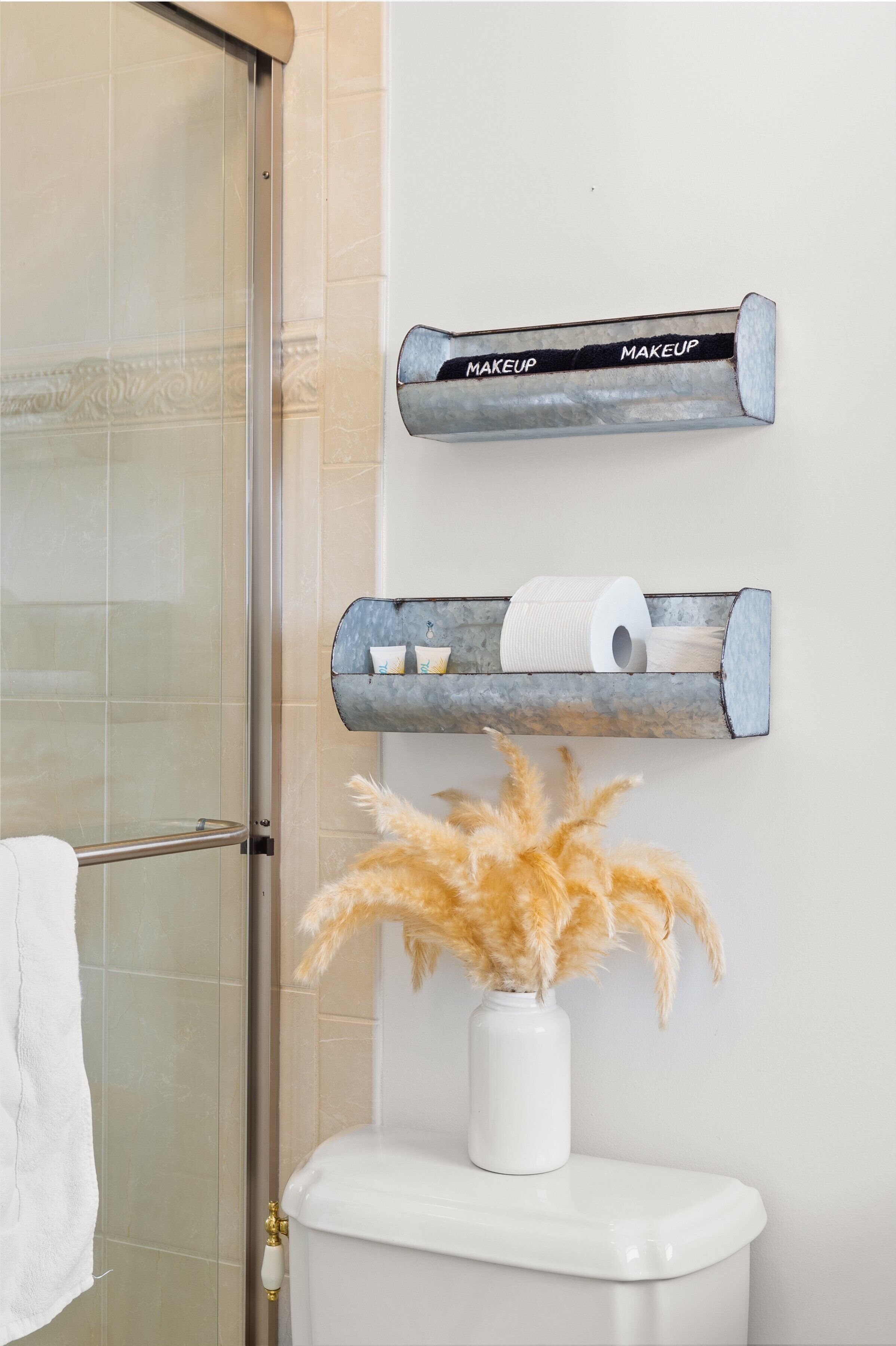 Un baño con puerta de ducha de vidrio, un inodoro con un jarrón blanco de flores secas y dos dispensadores metálicos etiquetados como 