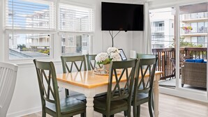 Een eetgedeelte met een houten tafel en groene stoelen, een flatscreen televisie aan de muur en een balkon met rieten meubilair.