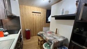 Cocina compacta con pared de madera, una pequeña mesa de comedor y una banca. Se observa un bote de basura rojo y un estante negro con un rollo de toallas de papel.
