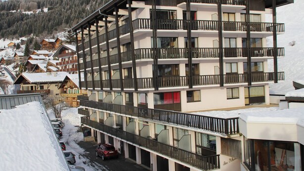 Un hotel de varios pisos con balcones, un estacionamiento y una montaña nevada al fondo.