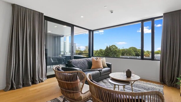 A modern living room with a sofa, two wicker chairs, a coffee table, and large windows offering a view of greenery and buildings.