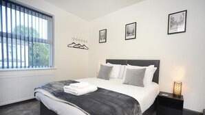 A bedroom with a bed, a bedside table with a lamp, and a window with blinds. - Spacious home with 8 beds, perfect for work trips or family stays. Enjoy the comfort of free parking, fast Wi-Fi, and a fully equipped space to relax or get things done. (Southampton)