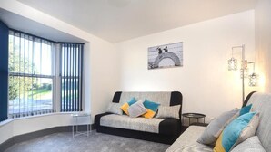 A modern living room with a sofa, a small table, and a large window with blinds. - Spacious home with 8 beds, perfect for work trips or family stays. Enjoy the comfort of free parking, fast Wi-Fi, and a fully equipped space to relax or get things done. (Southampton)
