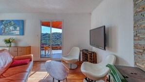 Un salon moderne avec un canapé en cuir, des fauteuils blancs, un téléviseur à écran plat et une grande fenêtre donnant sur un paysage verdoyant.