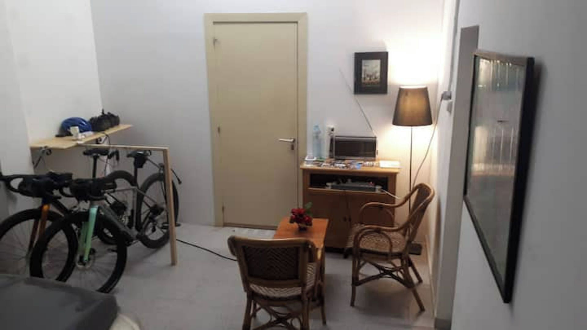 A small room with a door, a desk with a computer, a chair, a bicycle, and a lamp.