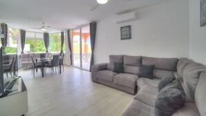 Ein geräumiger Wohnbereich mit Sofa, Esstisch und Stühlen. Ein Deckenventilator und ein Fenster mit Vorhängen sind vorhanden.
