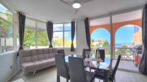 Ein modernes Wohnzimmer mit einer Couch, Esstisch und Stühlen, großen Fenstern mit Vorhängen und Blick auf das Meer und Gebäude.