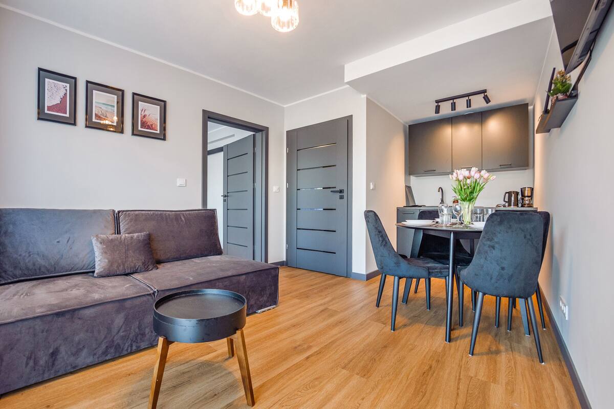A modern living room with a grey sofa, a small round table, and a dining area with a table set for two.
