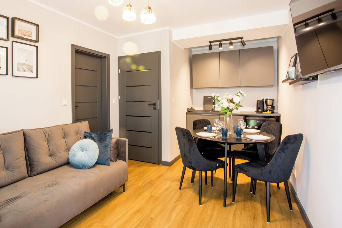 A modern living room with a brown sofa, a dining table with chairs, and a kitchen area with cabinets and a coffee machine.