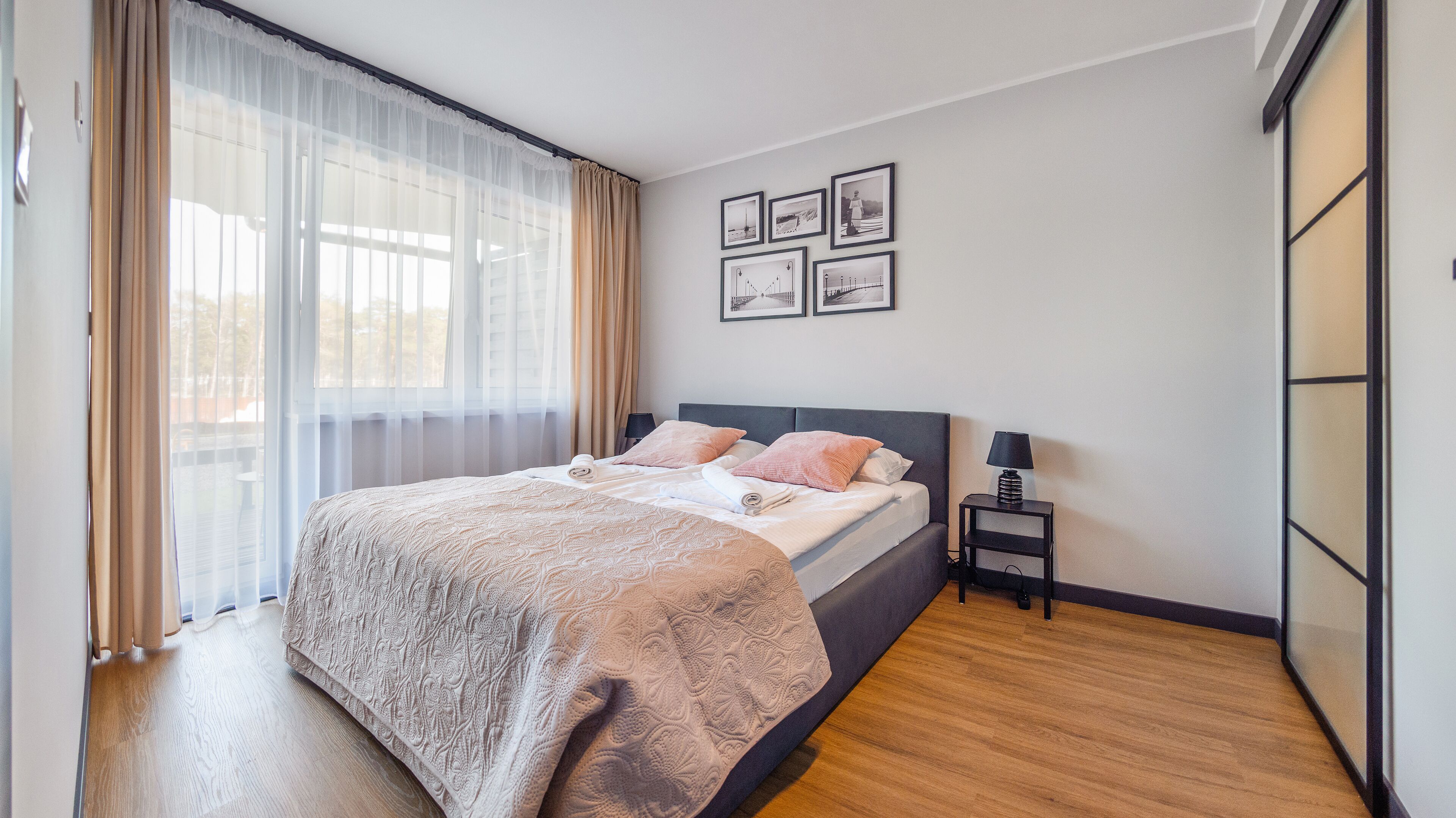 A neatly arranged bedroom with a large bed, a bedside table with a lamp, and a window with sheer curtains.
