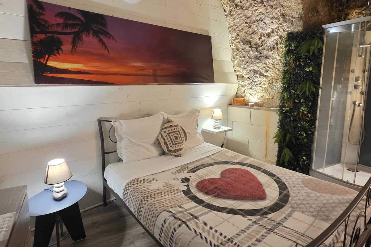 A bedroom with a bed, a nightstand, a lamp, a painting, a shower, and a wall with a textured surface.