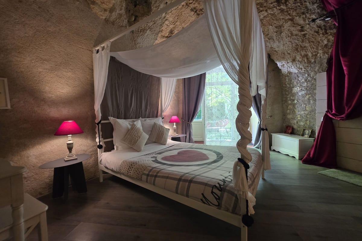 A four-poster canopy bed with a patterned bedspread, two bedside lamps, and a nightstand with a framed photo.