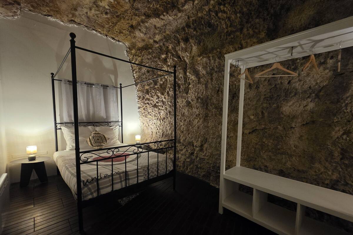 A four-poster bed with a canopy, bedside lamps, a white bedside table, and a white bench with shelves against a textured stone wall.