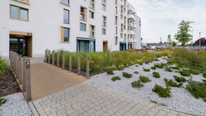 Un edificio moderno de varios pisos, entrada con portón y jardín ajardinado con grava y plantas pequeñas.