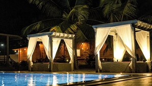 A nighttime pool area with two covered seating areas and palm trees in the background. - Brahmi Resort (Doddaballapur)