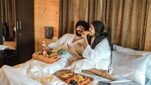 A couple in a cozy room with wooden walls, a bed with white linens, and a tray of fruits and snacks. - Brahmi Resort (Doddaballapur)