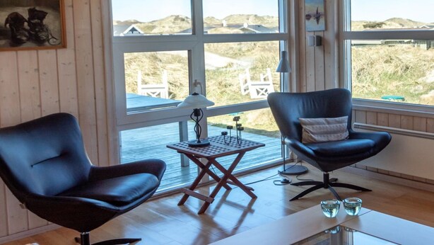 Une pièce avec deux fauteuils en cuir noir, une petite table en bois et une lampe. De grandes fenêtres offrent une vue sur la plage et les dunes.