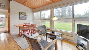 Un salón espacioso con techo de madera, ventanas amplias, una zona de comedor con sillas rojas, un sillón tejido gris, una mesita con una lámpara y un televisor sobre un mueble.