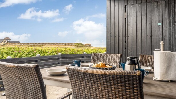 Un espace extérieur aménagé pour prendre les repas, avec des chaises en osier, une table sur laquelle se trouve une cafetière, et une vue sur le paysage.