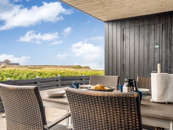 An outdoor dining area with wicker chairs, a table with a coffee pot, and a view of the landscape.