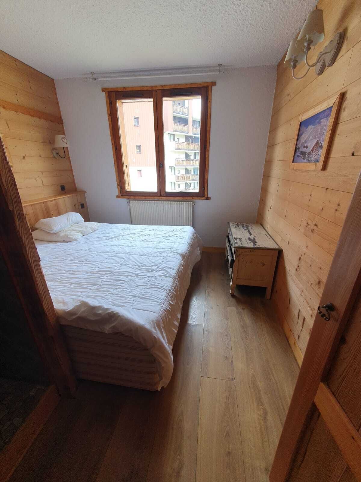Ein kleiner Raum mit Holzvertäfelung, einem Einzelbett, einem Heizkörper, einer kleinen Holzkiste und einem Fenster mit Blick auf ein Gebäude.