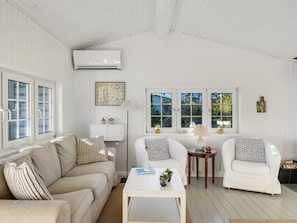 A room with a beige sofa, two white armchairs, a white coffee table, and a small side table with a lamp. There are two windows with white frames and a map on the wall.