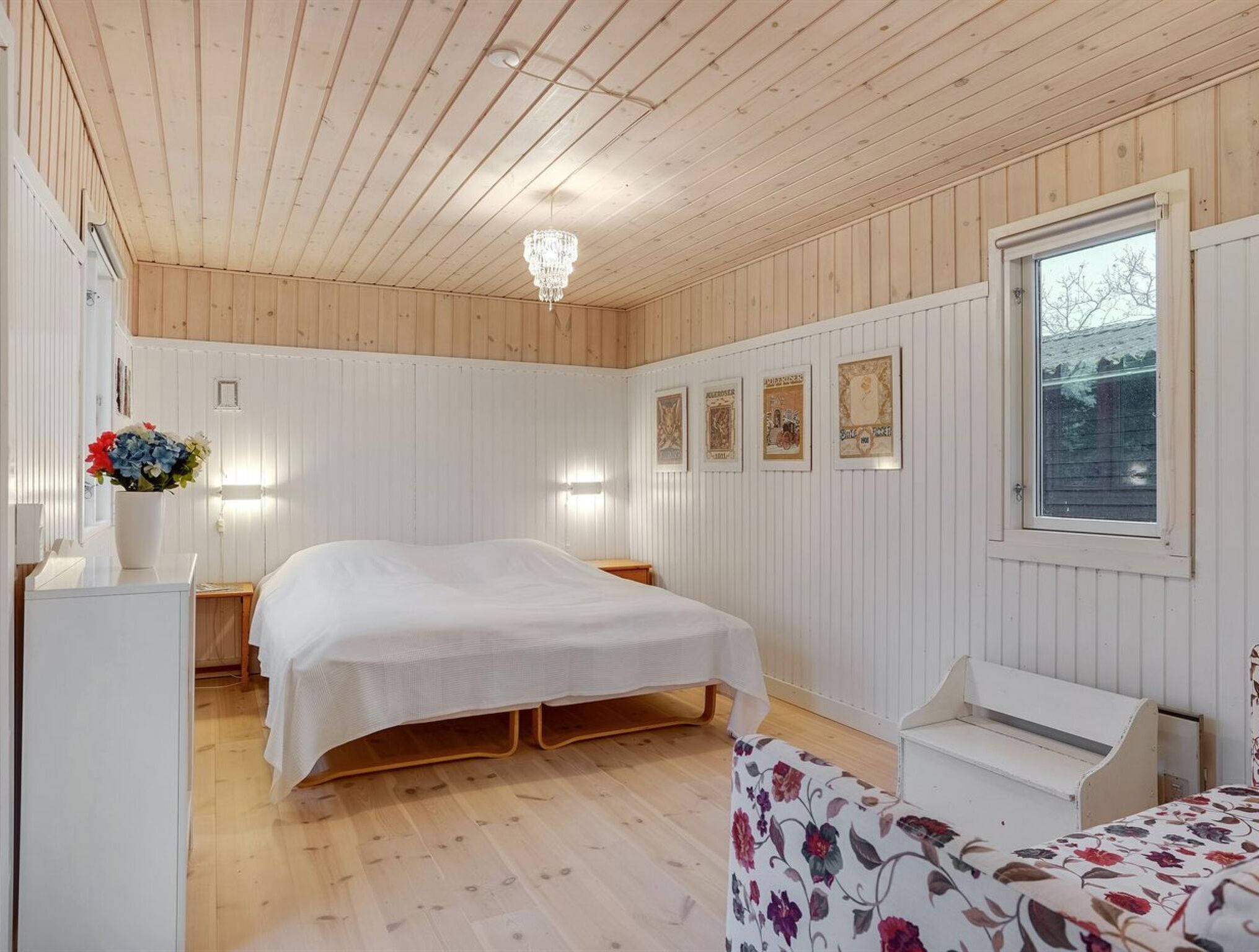 Un dormitorio acogedor con techo de madera, una cama con colcha blanca, una mesita de noche con un florero y una ventana con vistas a árboles.