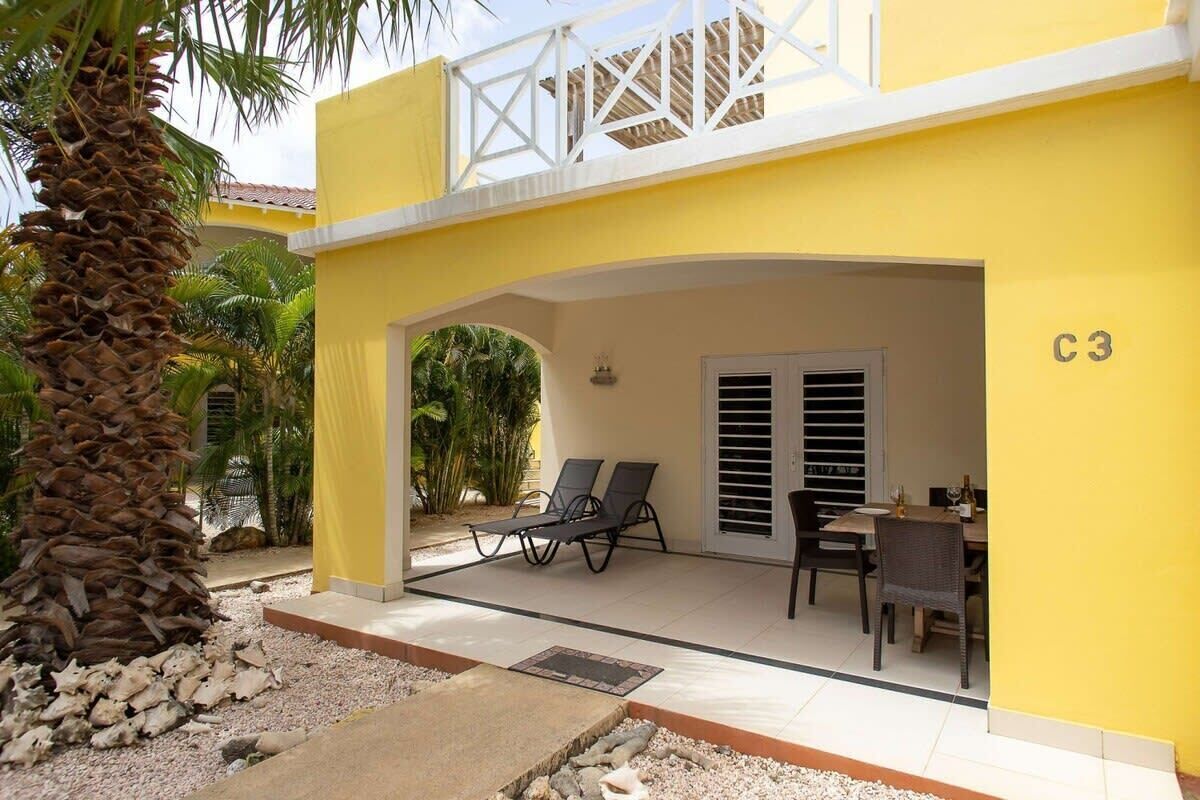 A yellow building with a covered patio area, numbered C3, featuring a table and chairs, and a palm tree.