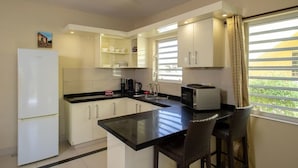 A modern kitchen with white cabinets, a black countertop, a refrigerator, a microwave, and a window with blinds. - Trupial D3 Relax & Dive Resort Bonaire Village (Kralendijk)