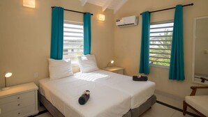 A bedroom with a bed, two bedside tables, a nightstand, a window with curtains, an air conditioning unit, and a mirror. - Prikichi C2 Relax & Dive Resort Bonaire Village (Kralendijk)