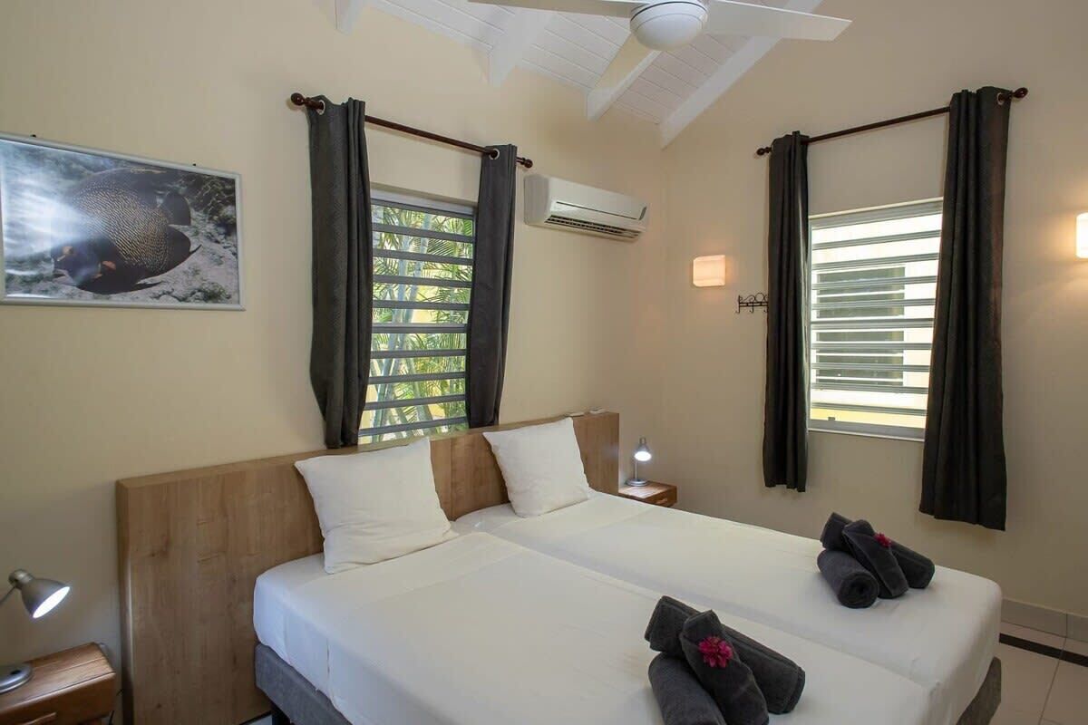 Cama doble con ropa de cama blanca, ventilador de techo, equipo de aire acondicionado, mesita de noche con lámpara y un cuadro en la pared.