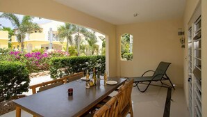 A dining area with a table and chairs, a lounge chair, and a view of a garden with palm trees and flowers. - Caracara I2 Relax & Dive Resort Bonaire Village (Kralendijk)