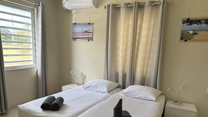 A hotel room with two beds, a window with blinds, a wall-mounted air conditioner, and a small bedside table with a lamp. - Caracara I2 Relax & Dive Resort Bonaire Village (Kralendijk)