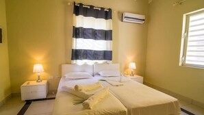 A hotel room with a bed, two bedside tables, a window with blinds, an air conditioning unit, and a lamp on each table. - Caracara J1 Relax & Dive Resort Bonaire Village (Kralendijk)