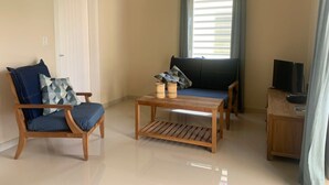 A living room with a wooden coffee table, a blue armchair, a sofa, and a television on a wooden cabinet. - Caracara J1 Relax & Dive Resort Bonaire Village (Kralendijk)