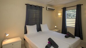 Una habitación de hotel con cama, mesita de noche, equipo de aire acondicionado, ventilador de techo y ventana con cortinas.
