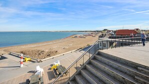 A wooden boardwalk leading to a beach with a clear view of the sea and distant buildings.