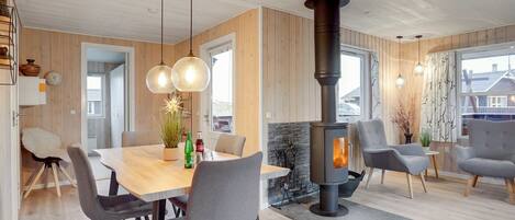 Ett modernt vardagsrum med en centralt placerad öppen spis, träpanel på väggarna, en matplats med stolar och hängande lampor.