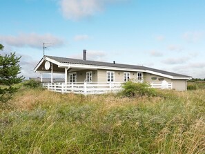 Une maison de plain-pied avec un porche, entourée d’herbes hautes et sous un ciel dégagé.
